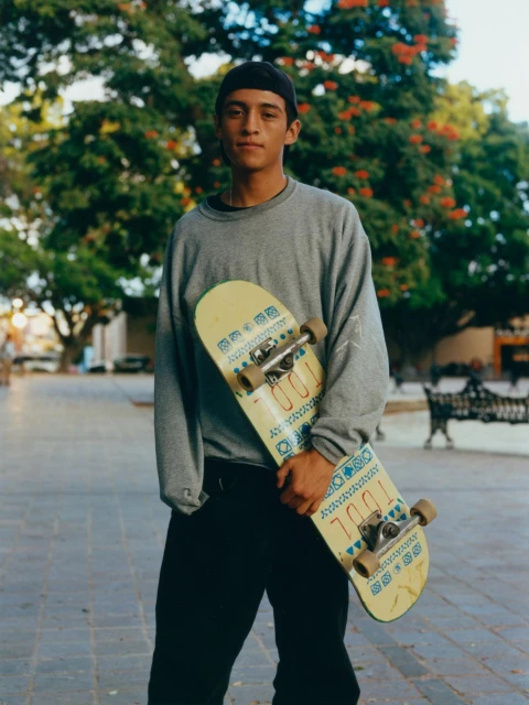 Young man in a black cap stands outside in a grey sweatshirt holding a skateboard with green trees in the background.
