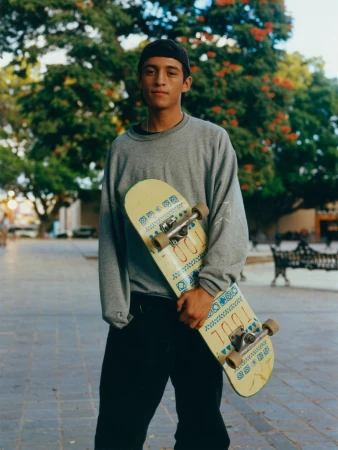 Young man in a black cap stands outside in a grey sweatshirt holding a skateboard with green trees in the background.
