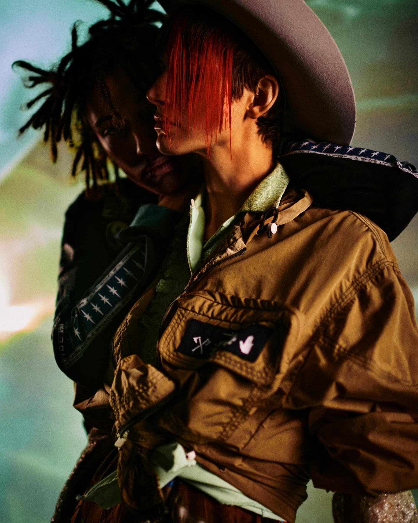 Two people posing intimately with warm lighting, one wearing a brown utility jacket and grey hat with red hair fringe