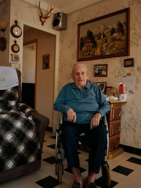 Elderly man sits in a wheelchair in an indoor living room with floral wallpaper and vintage decor; painting and antlers hang on the wall.