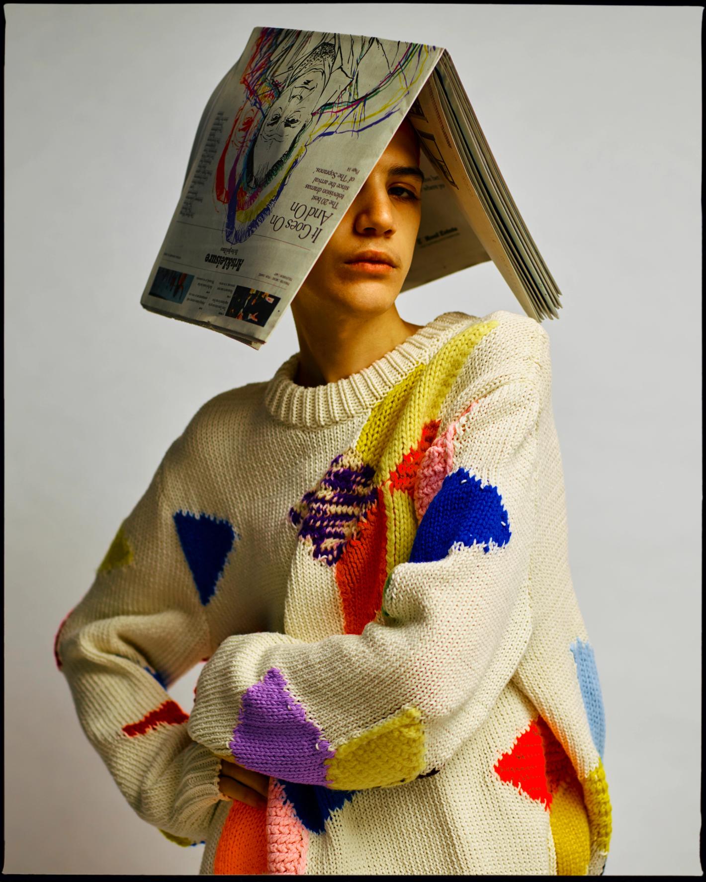 Portrait of a young adult wearing a patterned sweater and newspaper hat, looking at the camera against a neutral background.