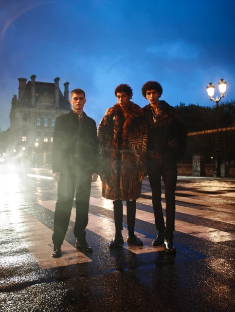 Three stylish adult male models posing outdoors at night on a wet city street with a historical building in the background under blue sky.