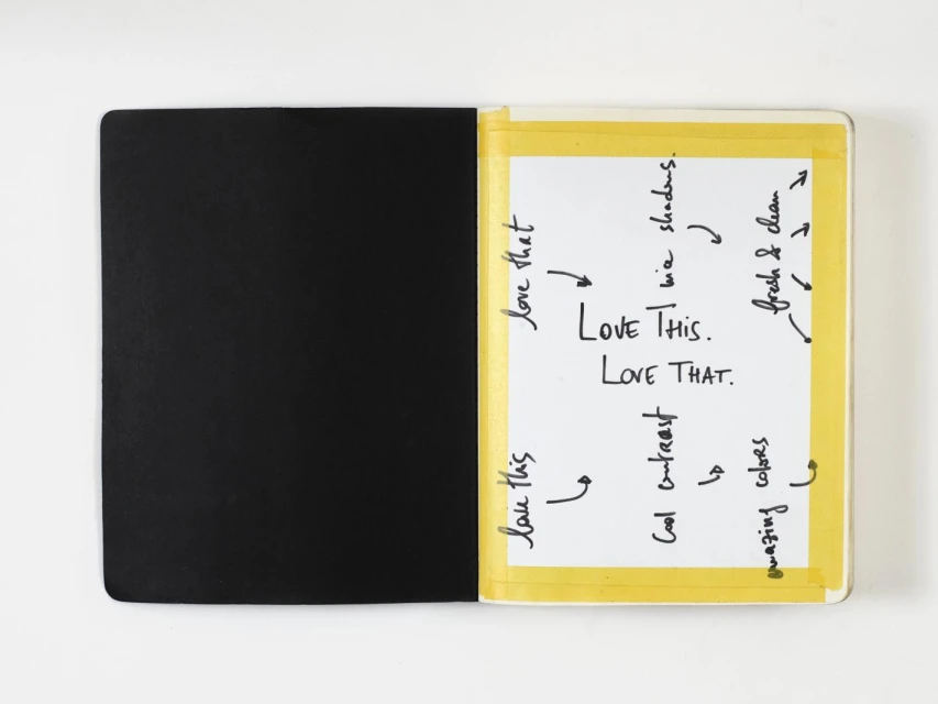 Open notebook with one black page and one white page with handwritten notes taped with yellow tape on a white surface.