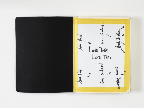 Open notebook with one black page and one white page with handwritten notes taped with yellow tape on a white surface.