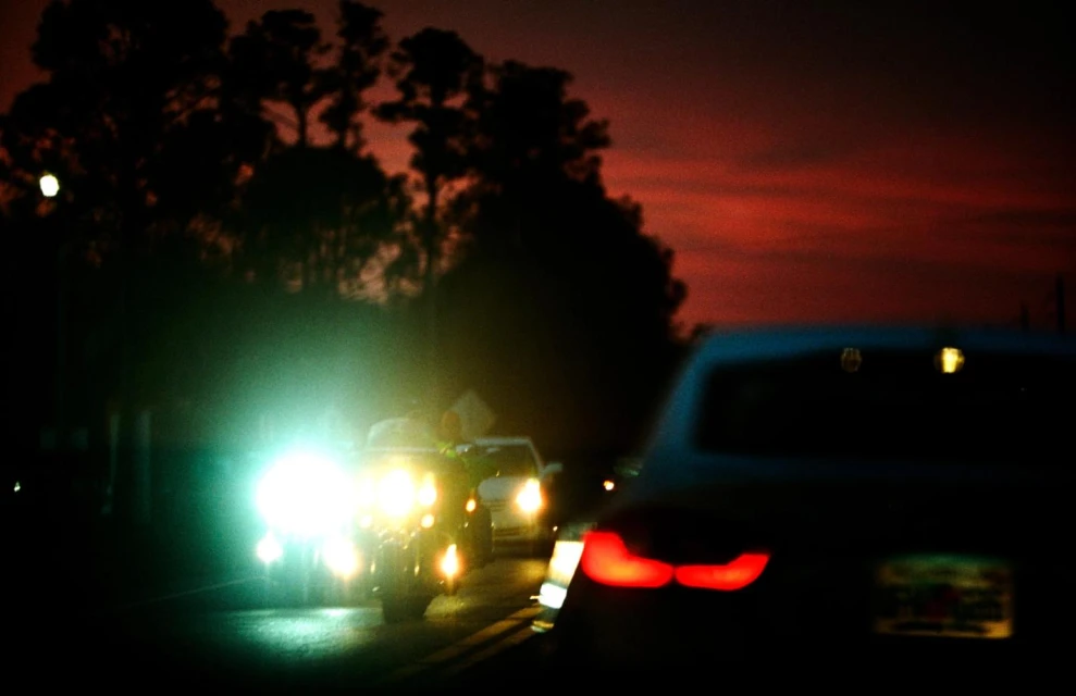 Multiple cars are on the road in the dark, with illuminated headlights and taillights visible against a twilight sky with silhouetted trees.