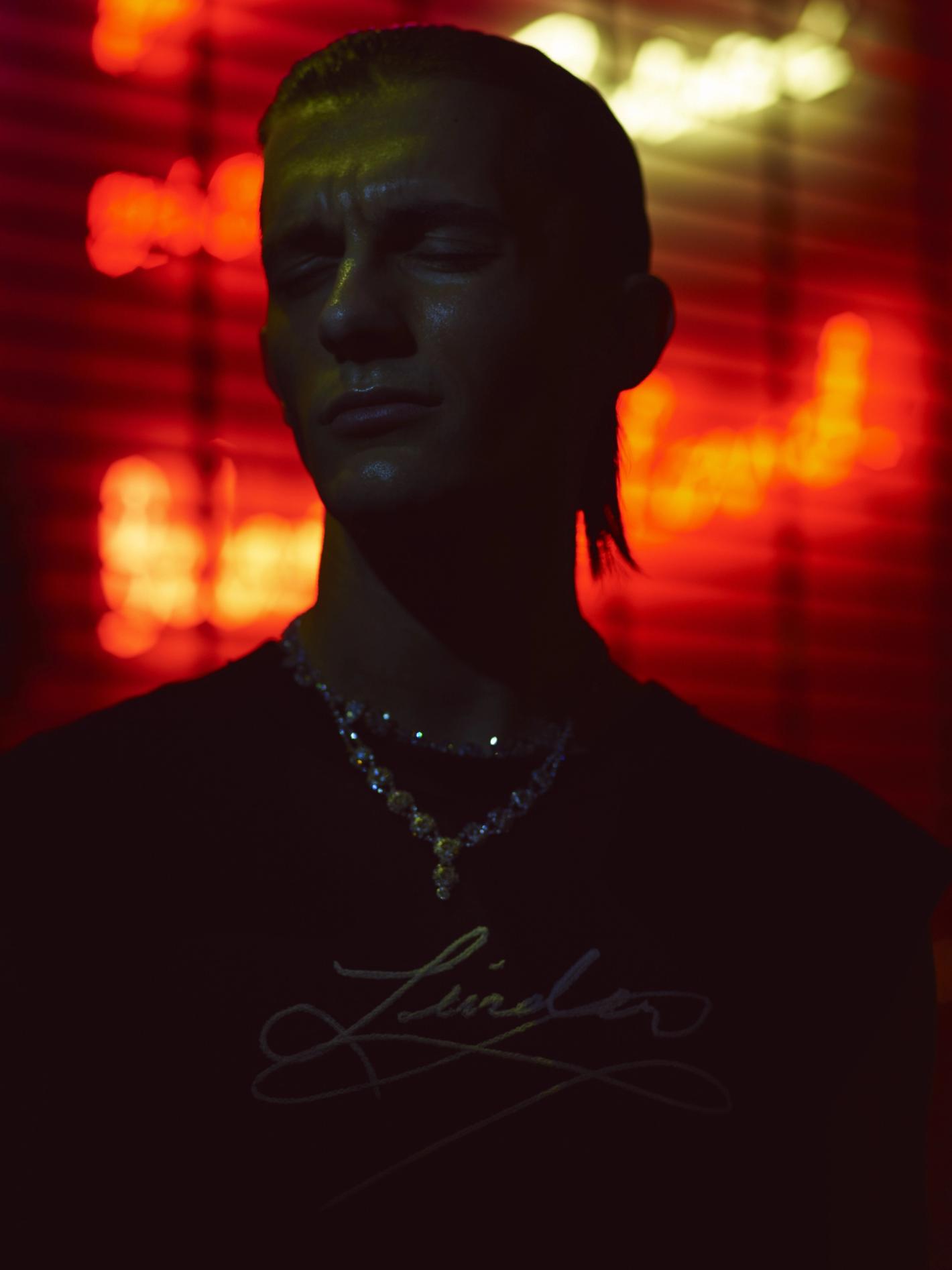 Portrait of a young adult man with slicked-back hair and a designer t-shirt, looking downward, illuminated by red and yellow neon lighting.