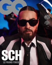 Close-up portrait of French rapper SCH wearing sunglasses, a black jacket, white shirt, and black tie against a motorcycle background
