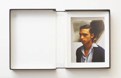 An open black book resting on a white surface displaying a portrait of a man with a newspaper folded over his head.
