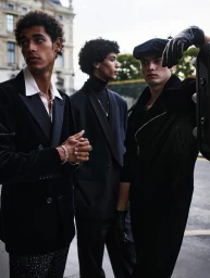 Three young men posing outdoors in stylish dark formal attire against an architectural background