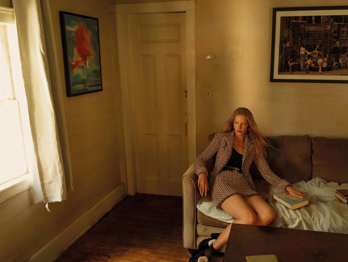 Woman in tweed skirt suit sitting on a couch in a warm-toned room with art, a table, and a window providing natural light.