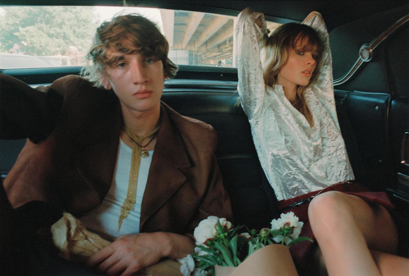 Two people in a vintage car, a young man in a brown jacket, and a woman in a white shirt, with a bouquet of white flowers.