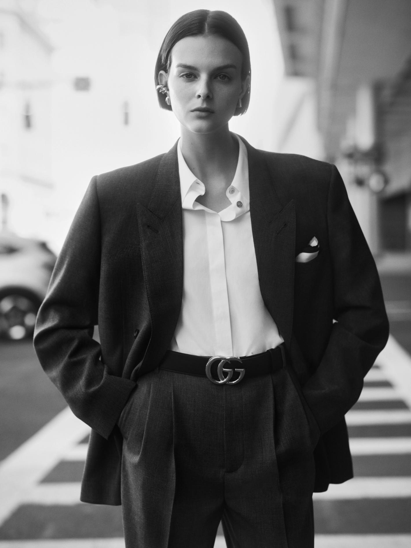 Elegant woman in a dark suit stands on a crosswalk with a blurred car in the background in this black and white fashion shot.