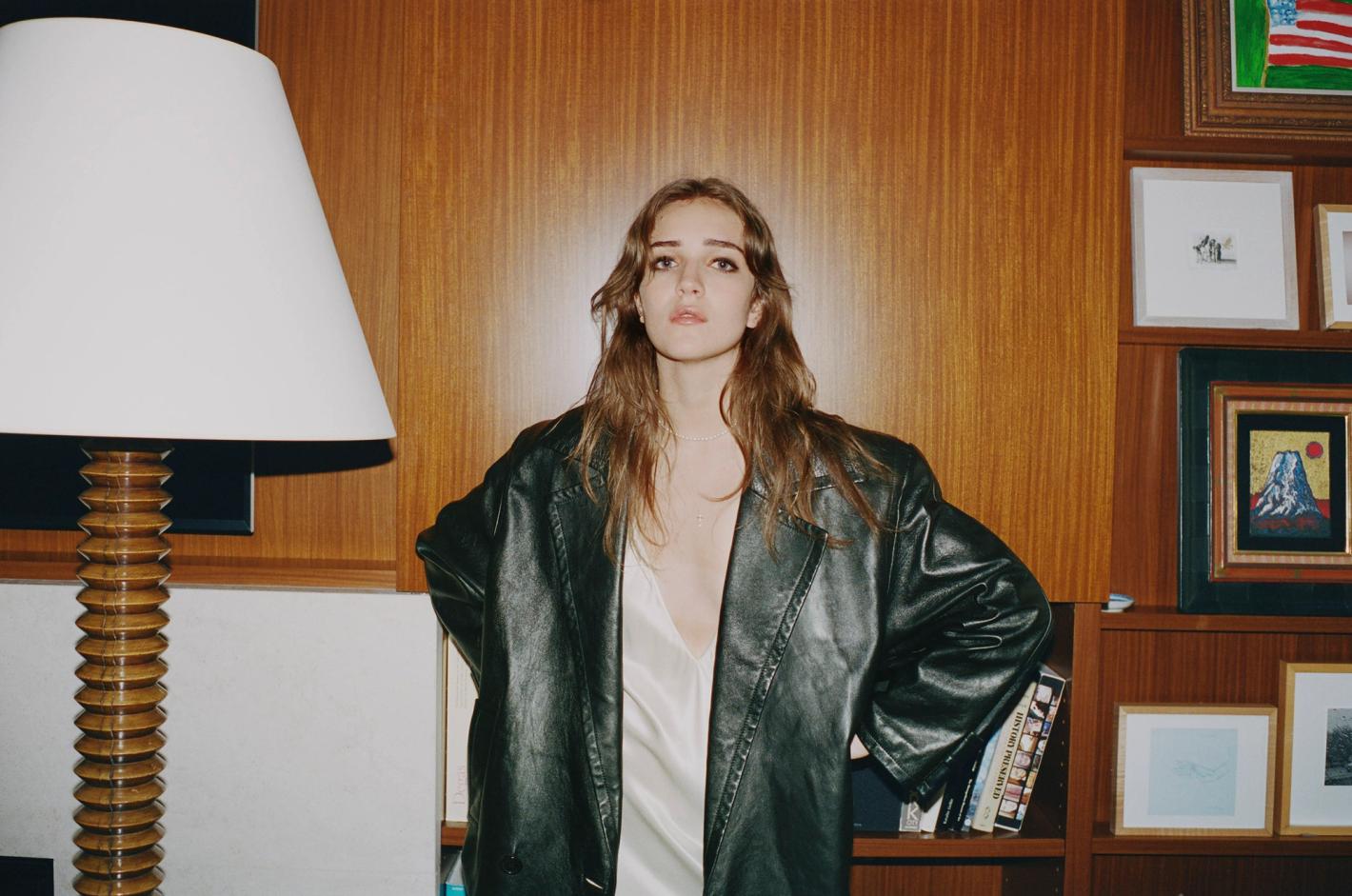 Portrait of a brunette woman in a black leather jacket, standing in an indoor setting with a wooden wall and books behind her.
