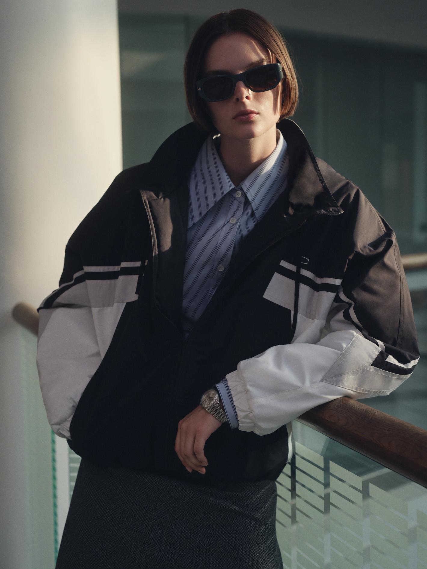 A young woman in a black and white jacket poses indoors, wearing sunglasses and a striped shirt, with a wristwatch visible on her left wrist.