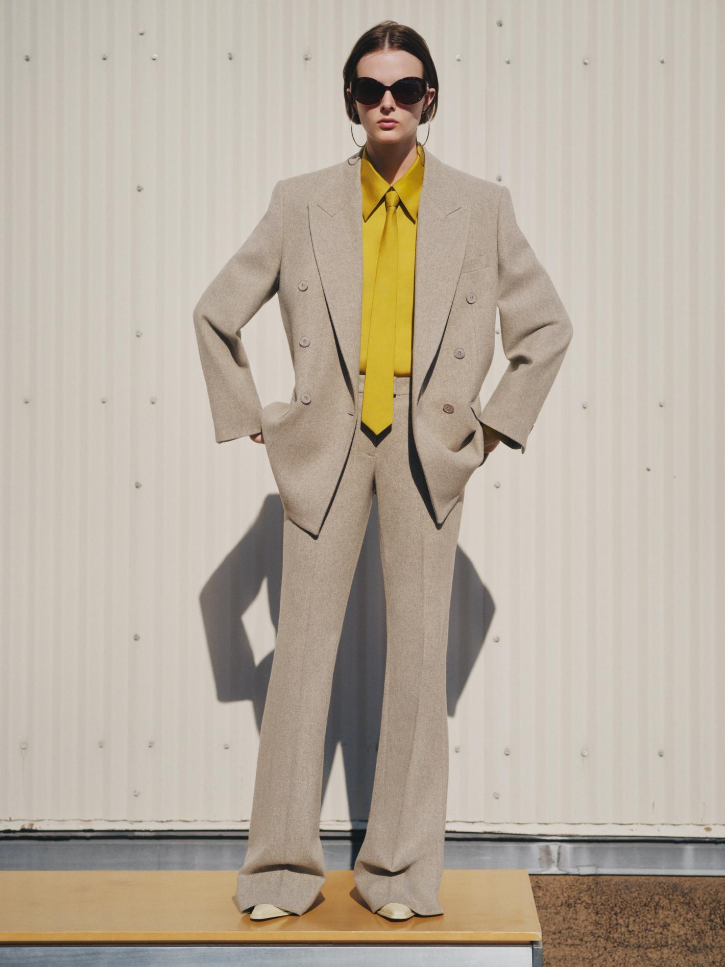 Full-length portrait of a stylish adult woman in a beige double-breasted suit with a yellow shirt and sunglasses, posing outdoors.