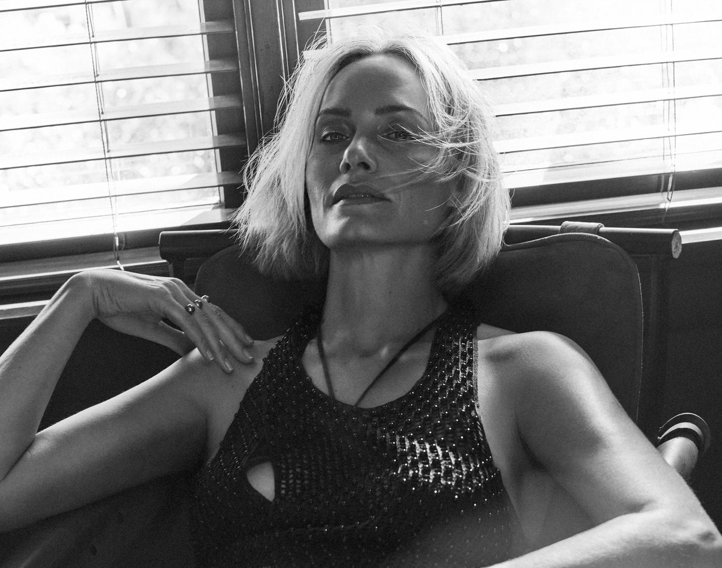 Black and white portrait of a fair-skinned adult woman with bob haircut, wearing a beaded top, seated in a chair by a window.