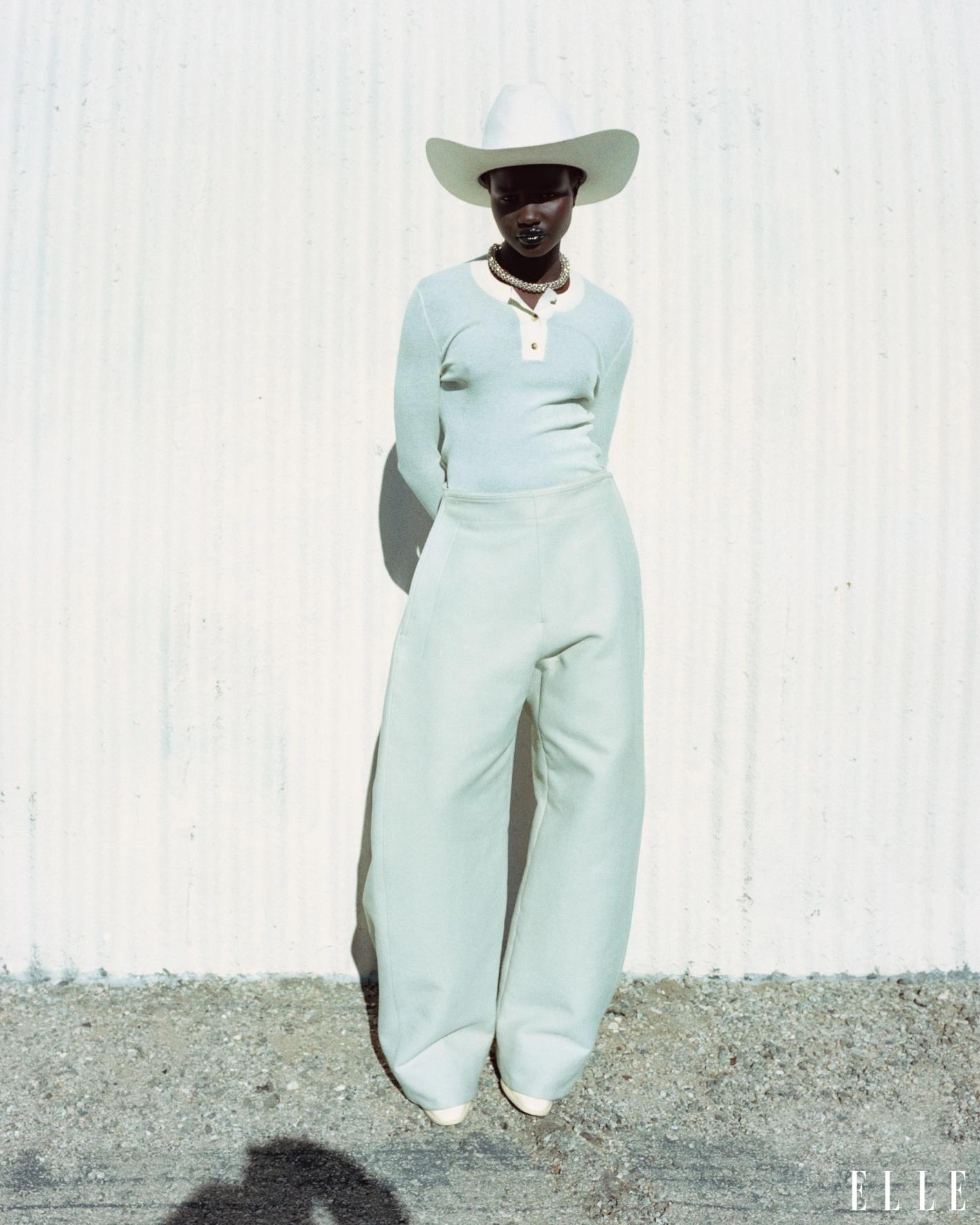 A slender woman with dark skin and a white cowboy hat poses in a light blue outfit against a textured white wall outdoors.