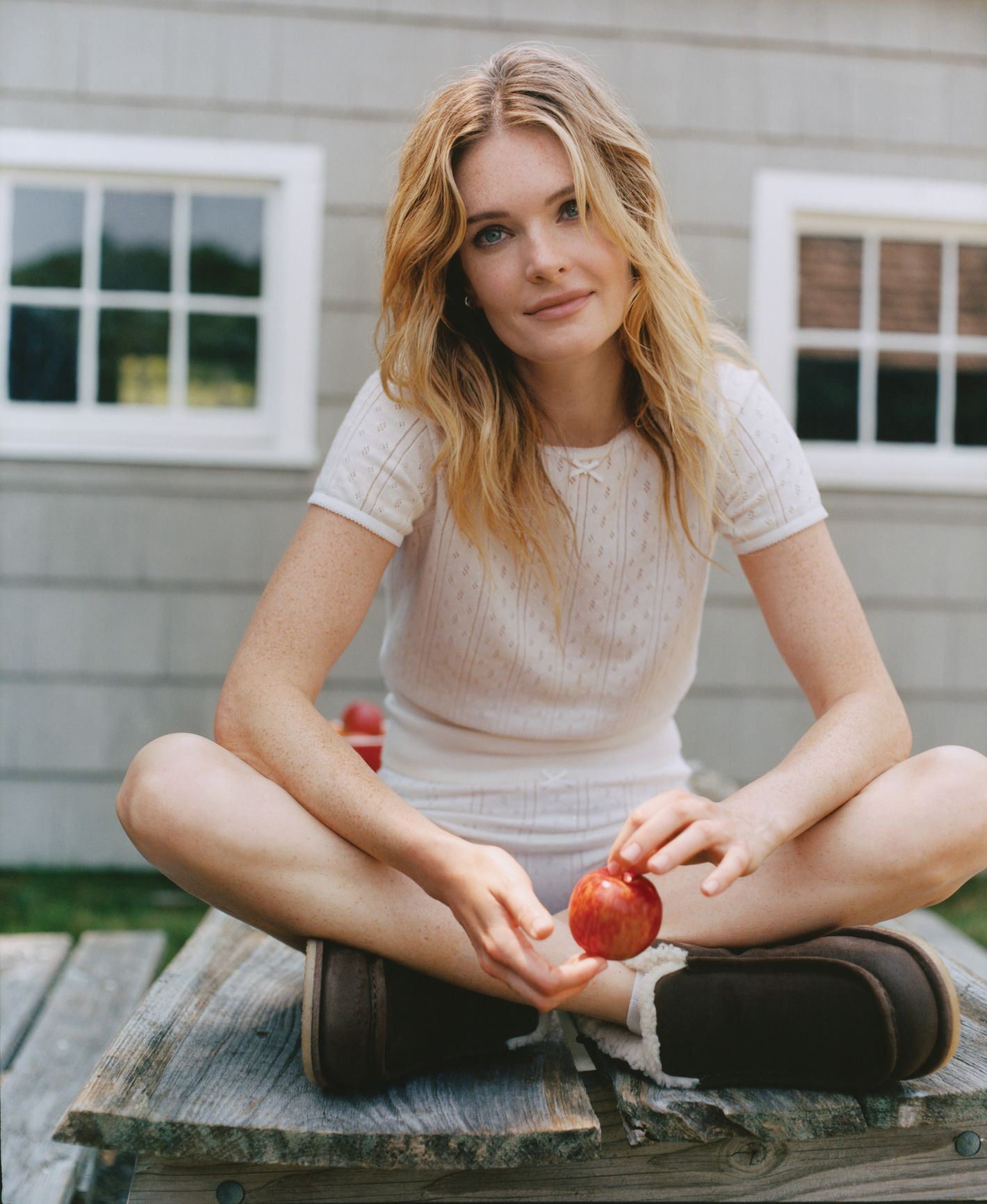 Blonde woman sits cross-legged on a picnic table holding an apple, outside with a grey house and white windows in the background.