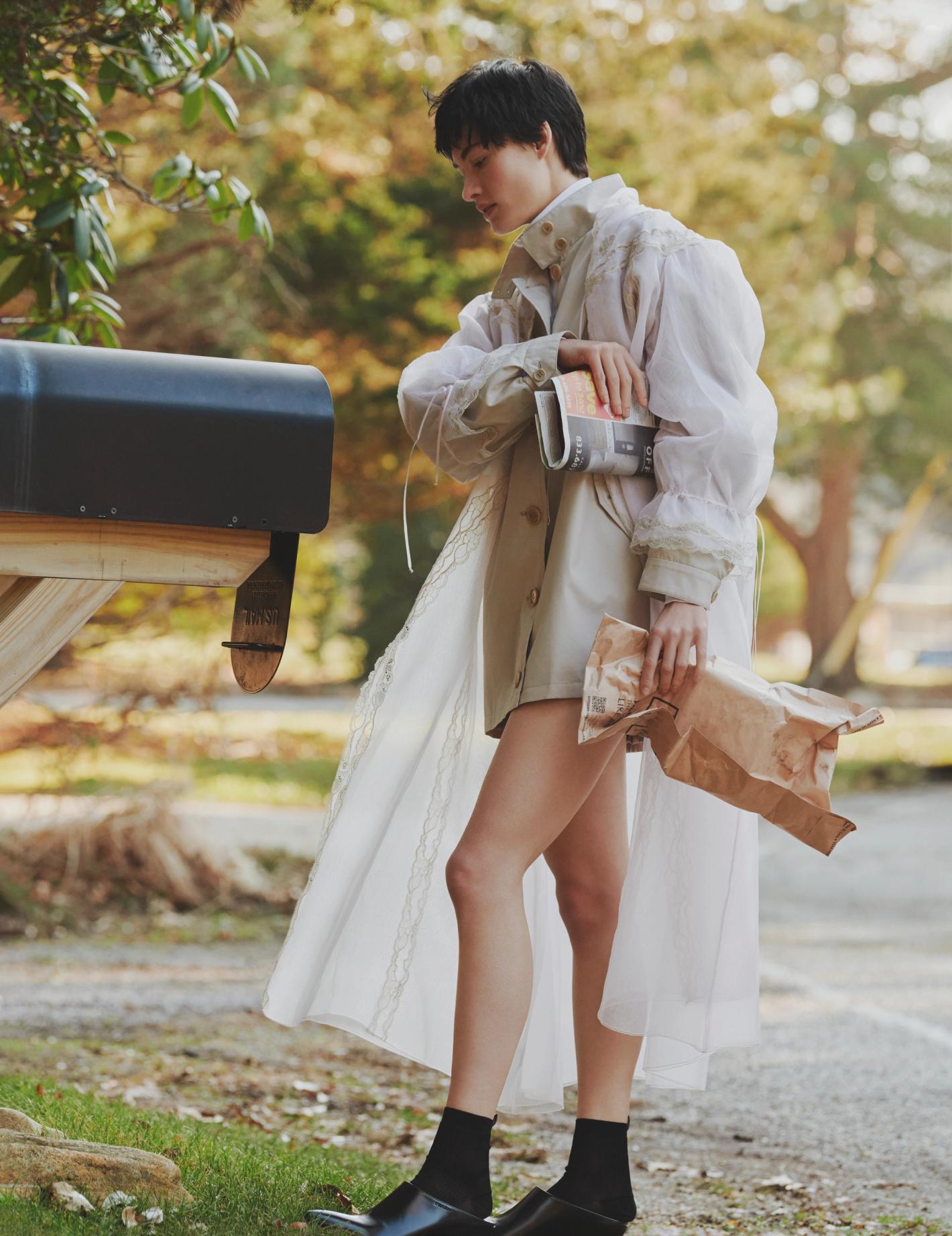 Woman in flowing trench coat stands near mailbox with newspaper and paper bag in rural setting with green trees and asphalt road.