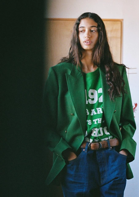 A woman with long brown hair models a casual look with a green blazer, graphic tee, and blue jeans in an indoor setting.