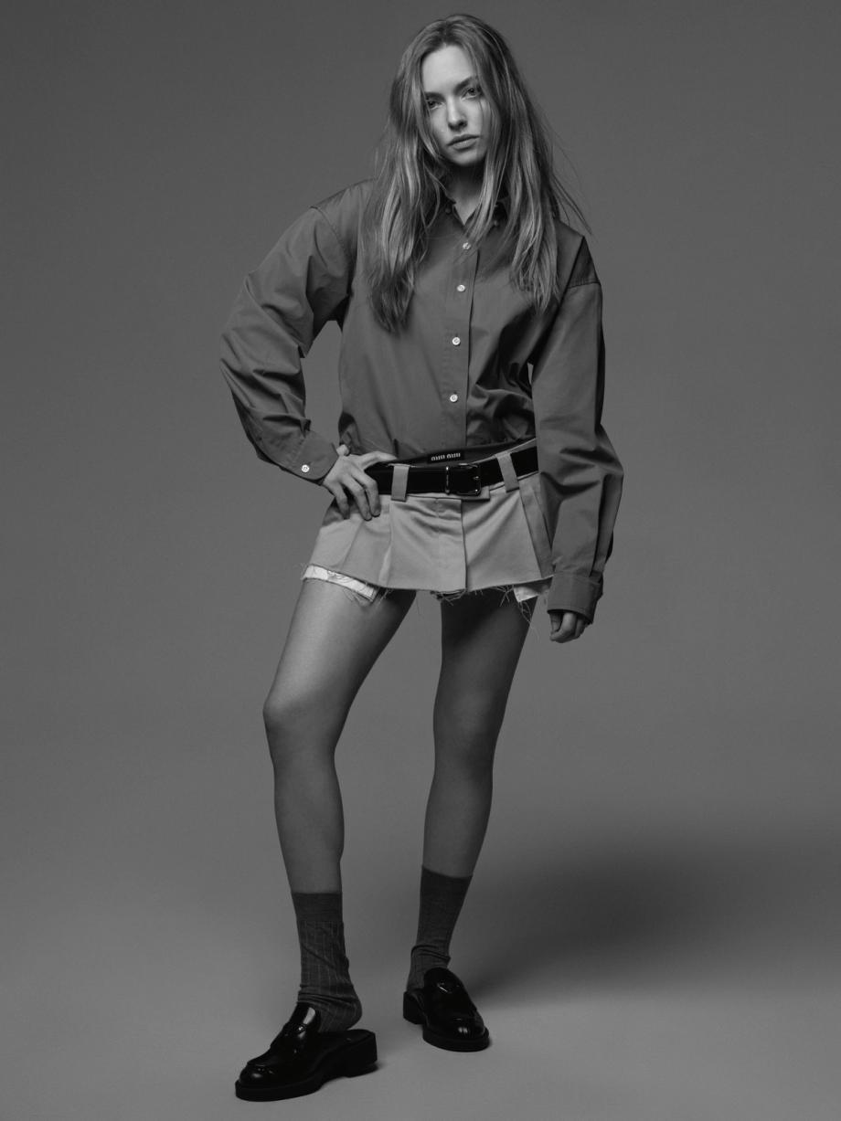 Full shot of an adult female model in an unbuttoned collared shirt and mini skirt, accessorized with a belt, socks, and loafers, in studio.