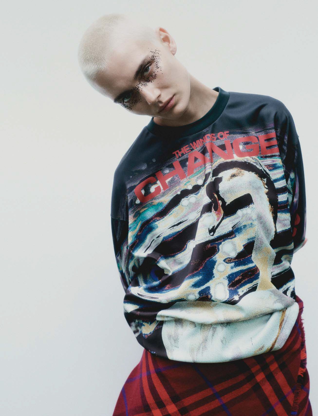Portrait of a young adult female with shaved head and glitter eyeshadow, wearing a graphic black shirt and plaid skirt against a white background.
