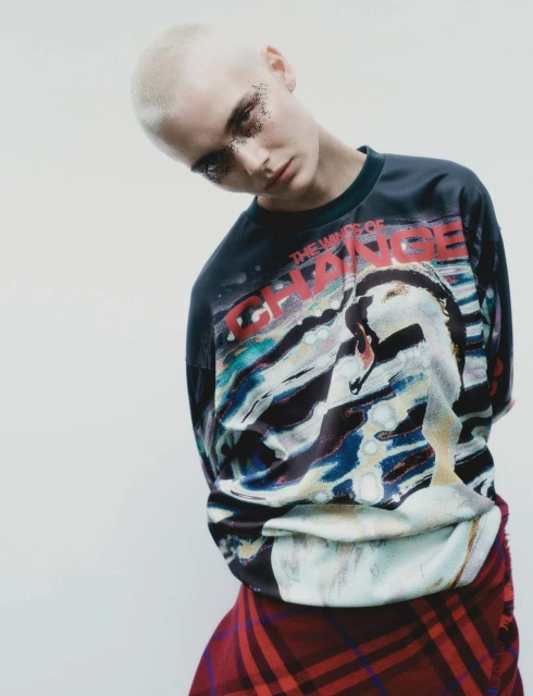 Portrait of a young adult female with shaved head and glitter eyeshadow, wearing a graphic black shirt and plaid skirt against a white background.