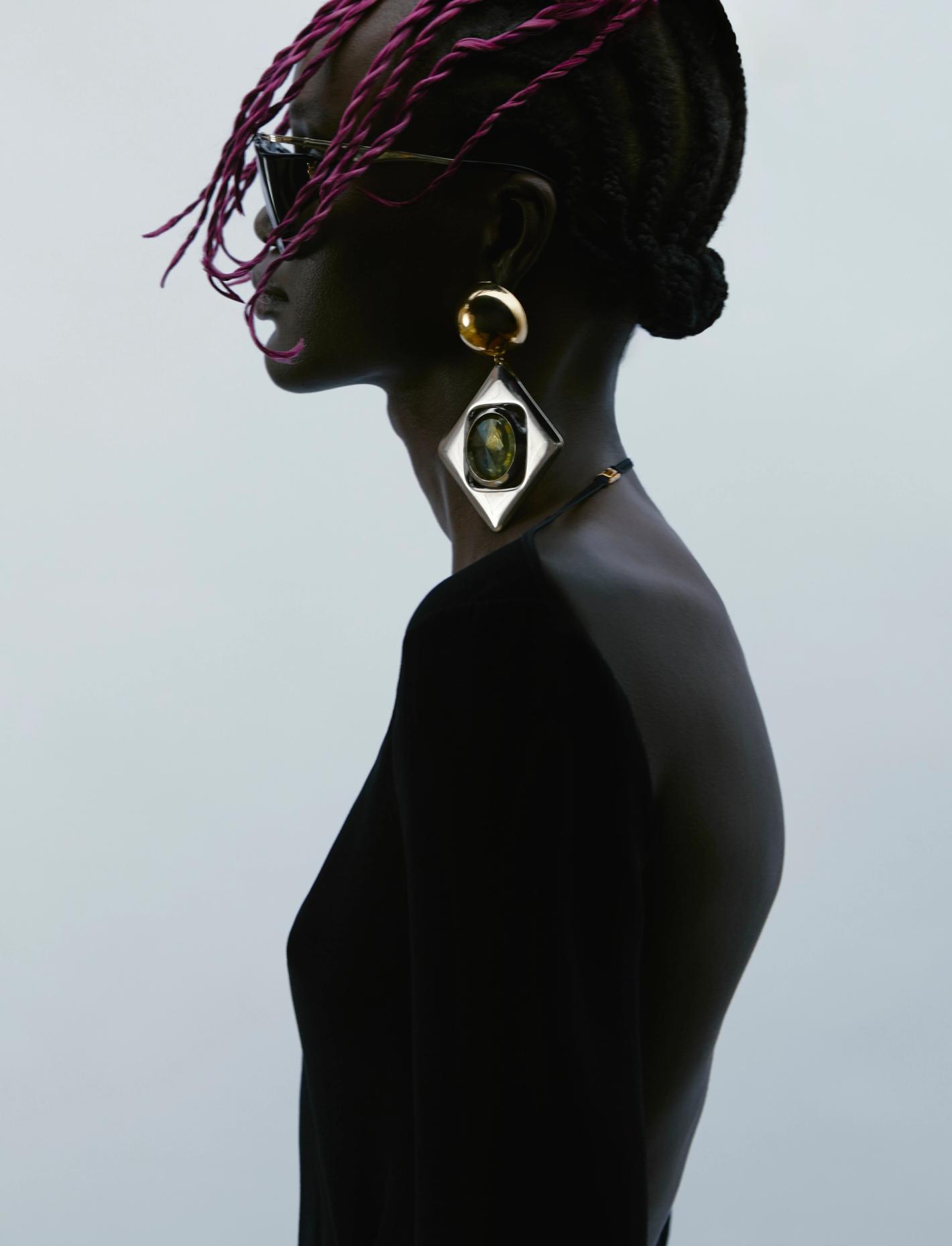 Profile shot of a woman with dark skin and pink braids, wearing a black dress and large gold earrings against a plain background.