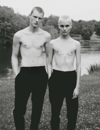 Monochrome portrait of two topless models with short hair wearing black trousers outdoors near a body of water.
