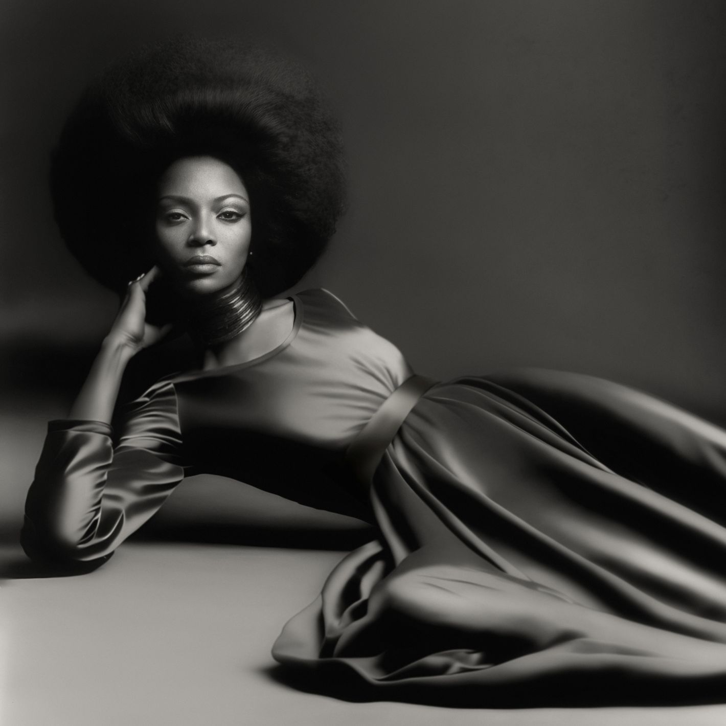 Black and white portrait of a woman with an afro wearing a long dress, lying on her side in a studio with a neutral background.