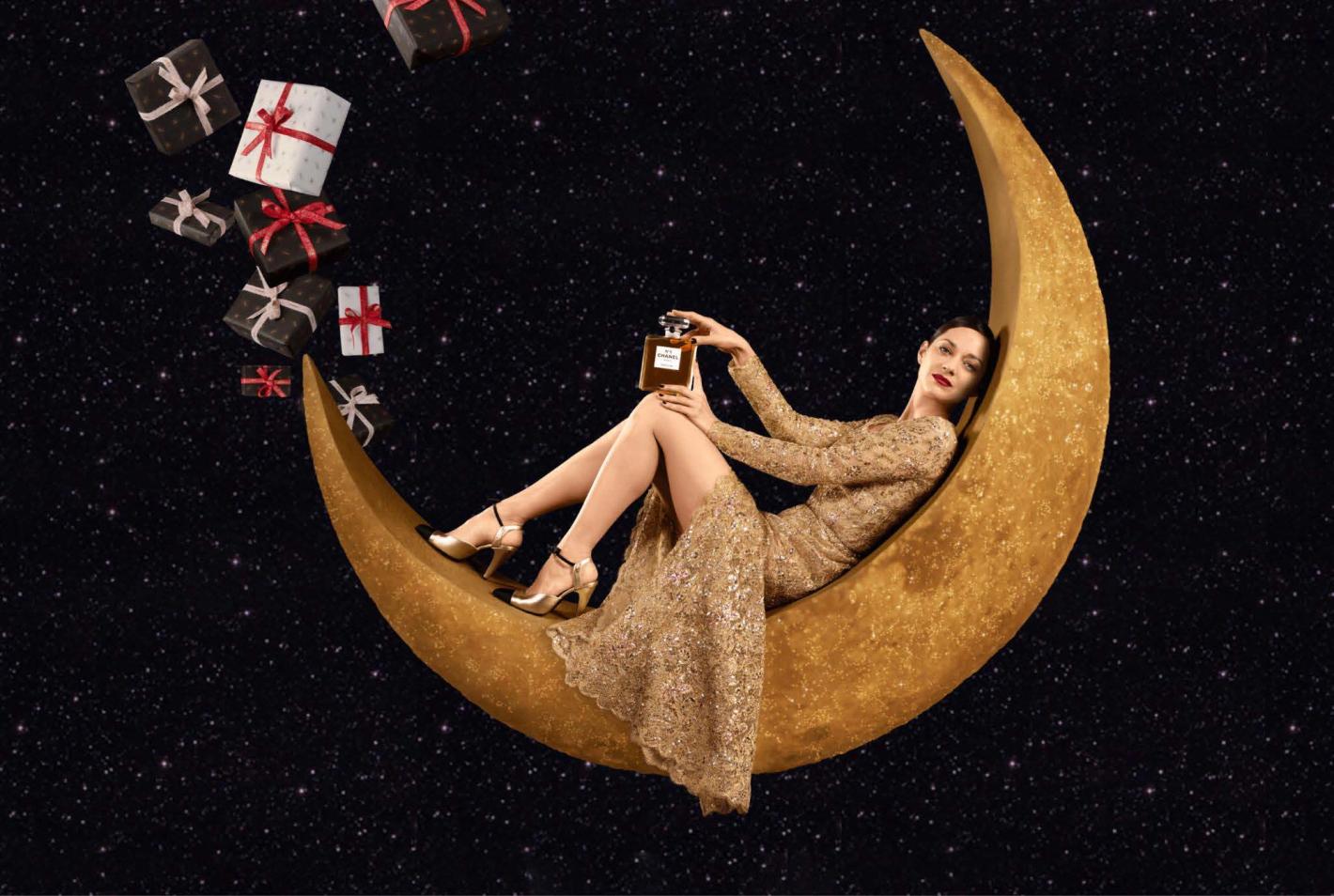 A woman in a gold gown reclines on a crescent moon, surrounded by floating gifts against a starry night backdrop, holding perfume.