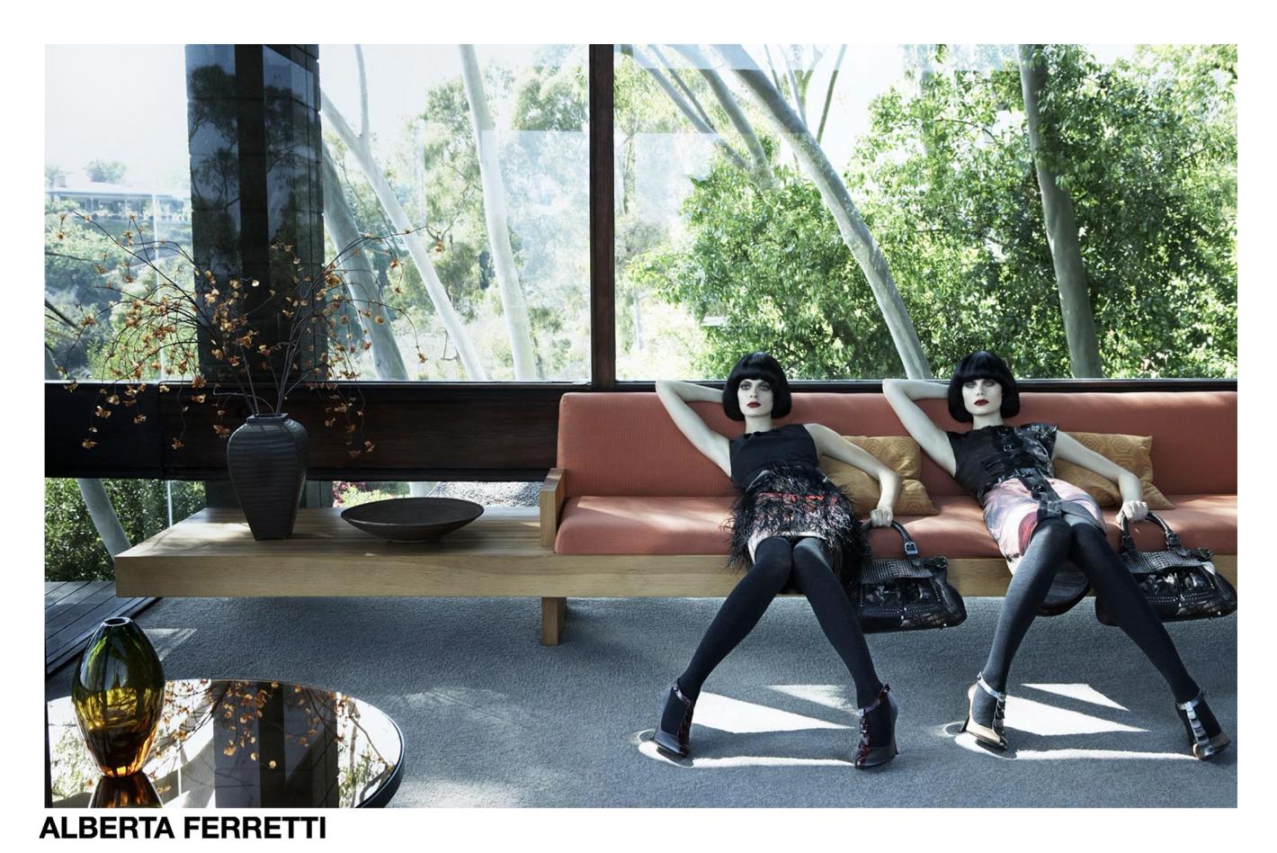 Two fashion models in chic dresses and heels lounge on an orange sofa in a modern home with large windows overlooking trees and greenery.