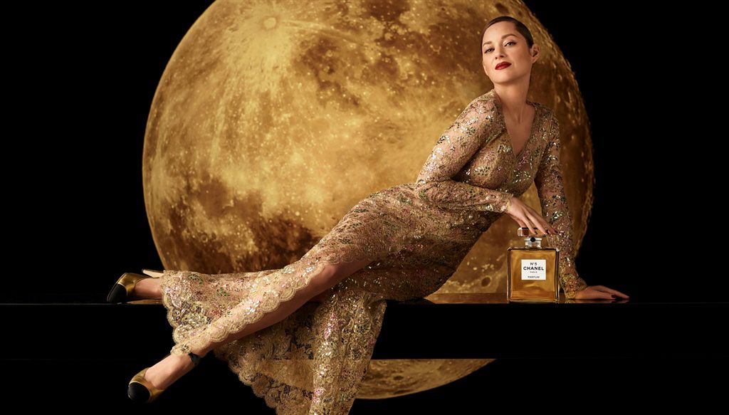 Elegant woman in a gold dress reclines with a Chanel perfume bottle against a lunar backdrop, showcasing beauty and fashion.