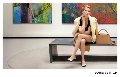 Léa Seydoux seated on a bench wearing a cream coat and black top with a tan handbag, in a gallery.