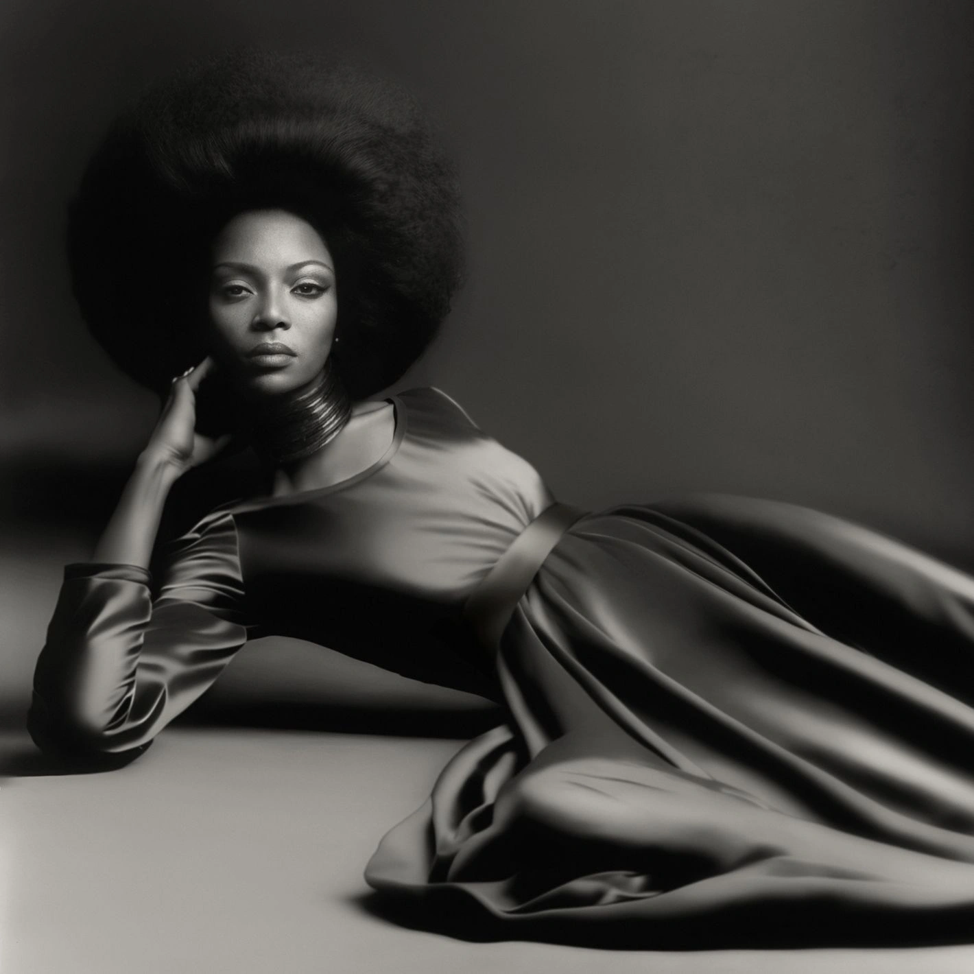 Black and white portrait of Naomi Campbell with a large afro wearing a satin gown and neck ring, posed on a studio floor.