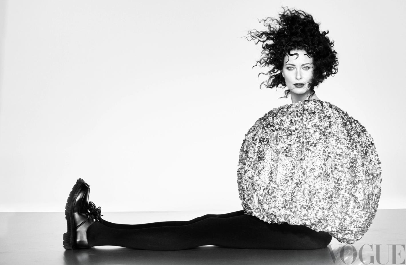 A woman with curly hair poses in a studio wearing a large, round, shimmering top, black leggings, and black shoes against a white backdrop.