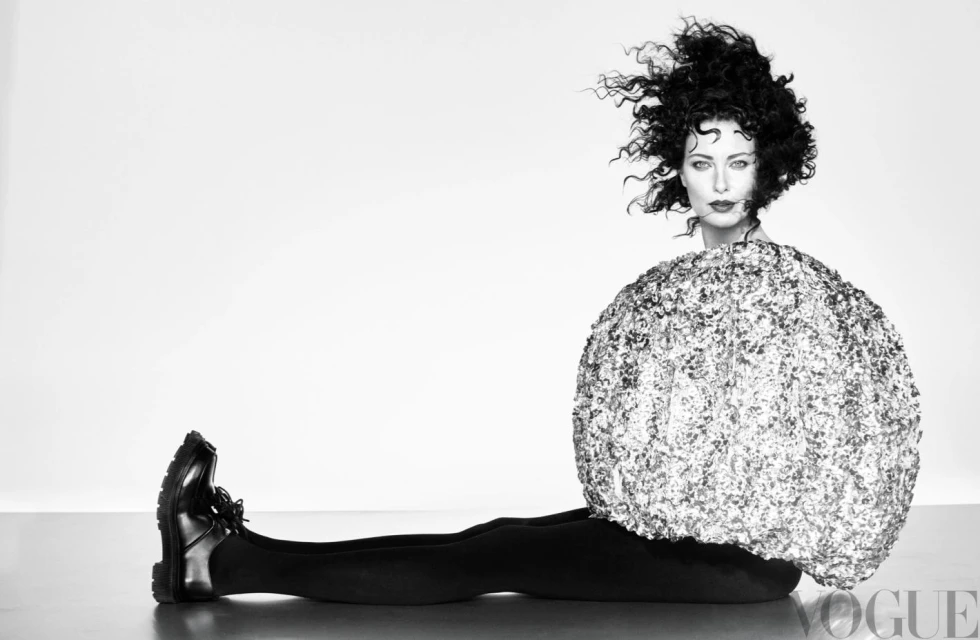 A woman with curly hair poses in a studio wearing a large, round, shimmering top, black leggings, and black shoes against a white backdrop.