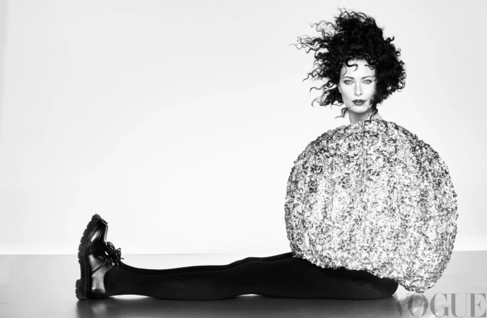 A woman with curly hair poses in a studio wearing a large, round, shimmering top, black leggings, and black shoes against a white backdrop.