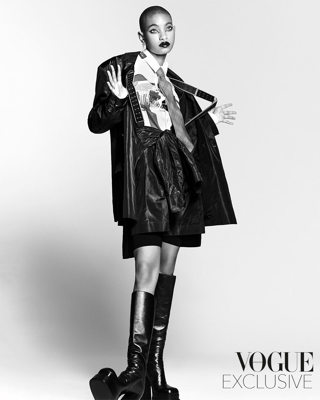 Monochrome fashion portrait of Willow Smith wearing leather coat, patterned shirt, tie, and tall boots against a white backdrop
