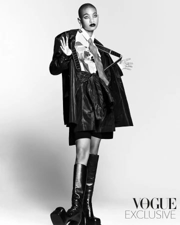 Monochrome fashion portrait of Willow Smith wearing leather coat, patterned shirt, tie, and tall boots against a white backdrop