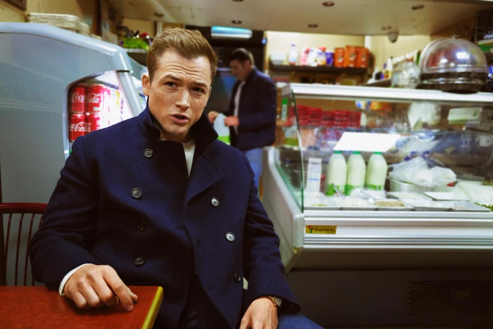 Adult male with light hair wears a navy pea coat seated at a table in a deli. Behind him, shelves of food and another person.