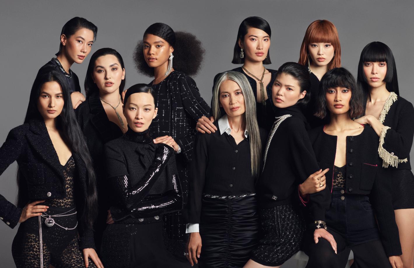Diverse group of ten adult women in fashionable black clothing, jewelry and elegant hairstyles, posing against a neutral grey backdrop.