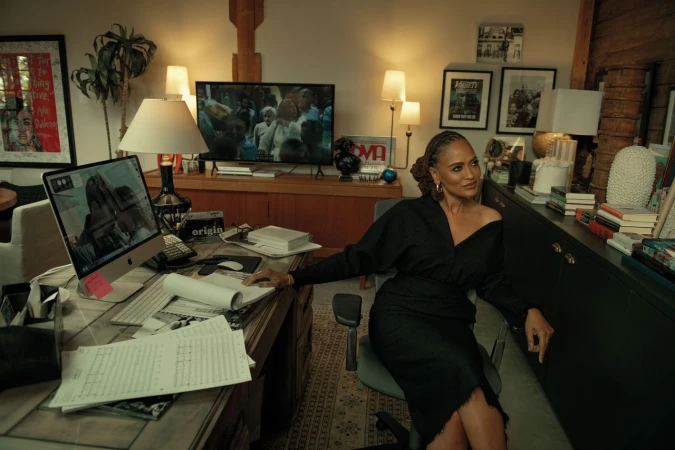 A woman in a black dress sits at a desk with a computer, open books, and papers in an office-like setting with decor and neutral colors.