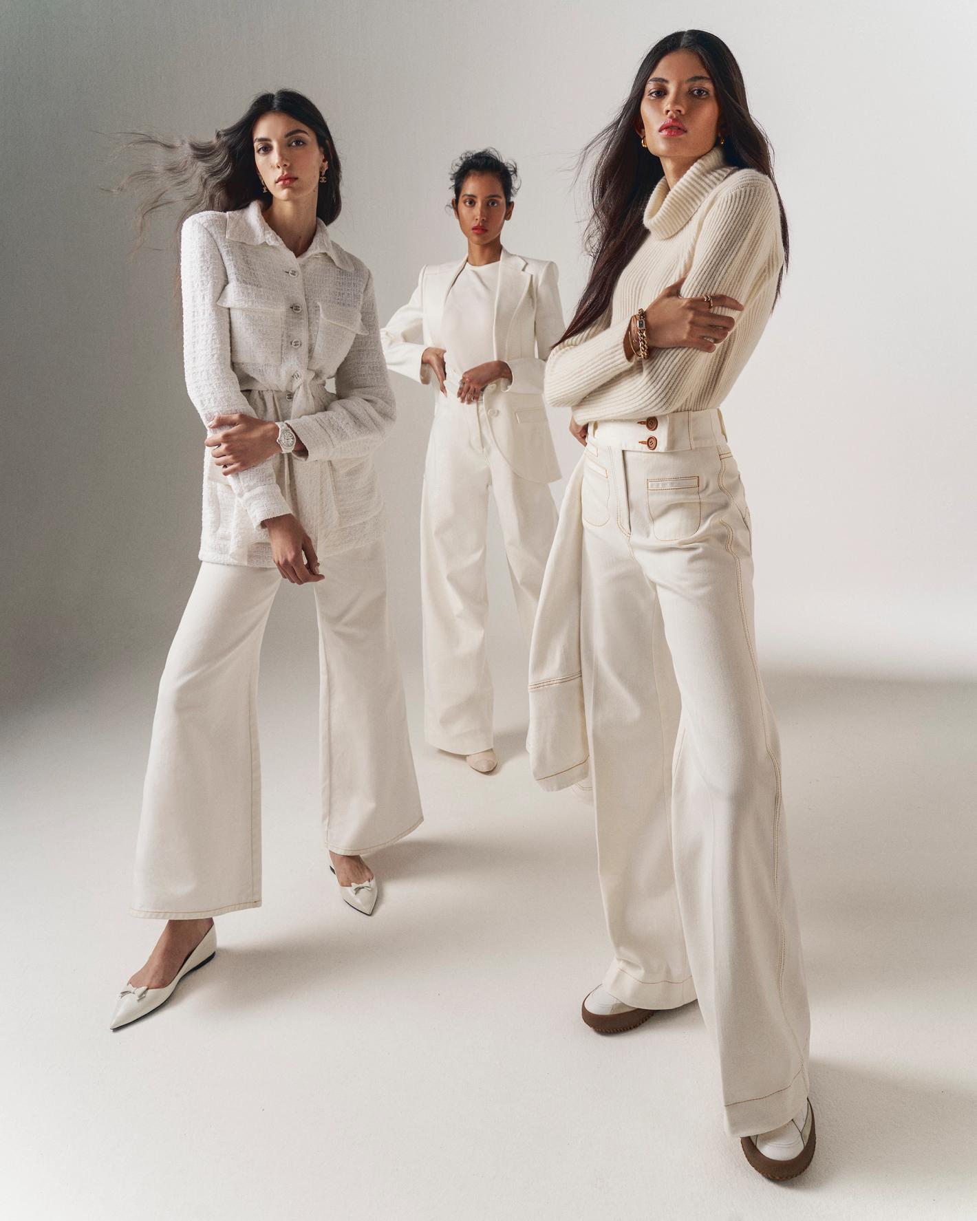 Three women pose in white outfits in a studio, one in a jacket and pants, one in a blazer and pants, and one in a sweater and pants.