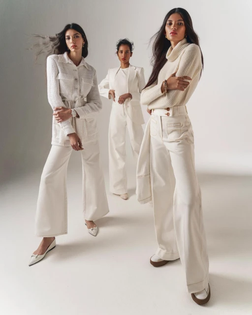 Three women pose in white outfits in a studio, one in a jacket and pants, one in a blazer and pants, and one in a sweater and pants.