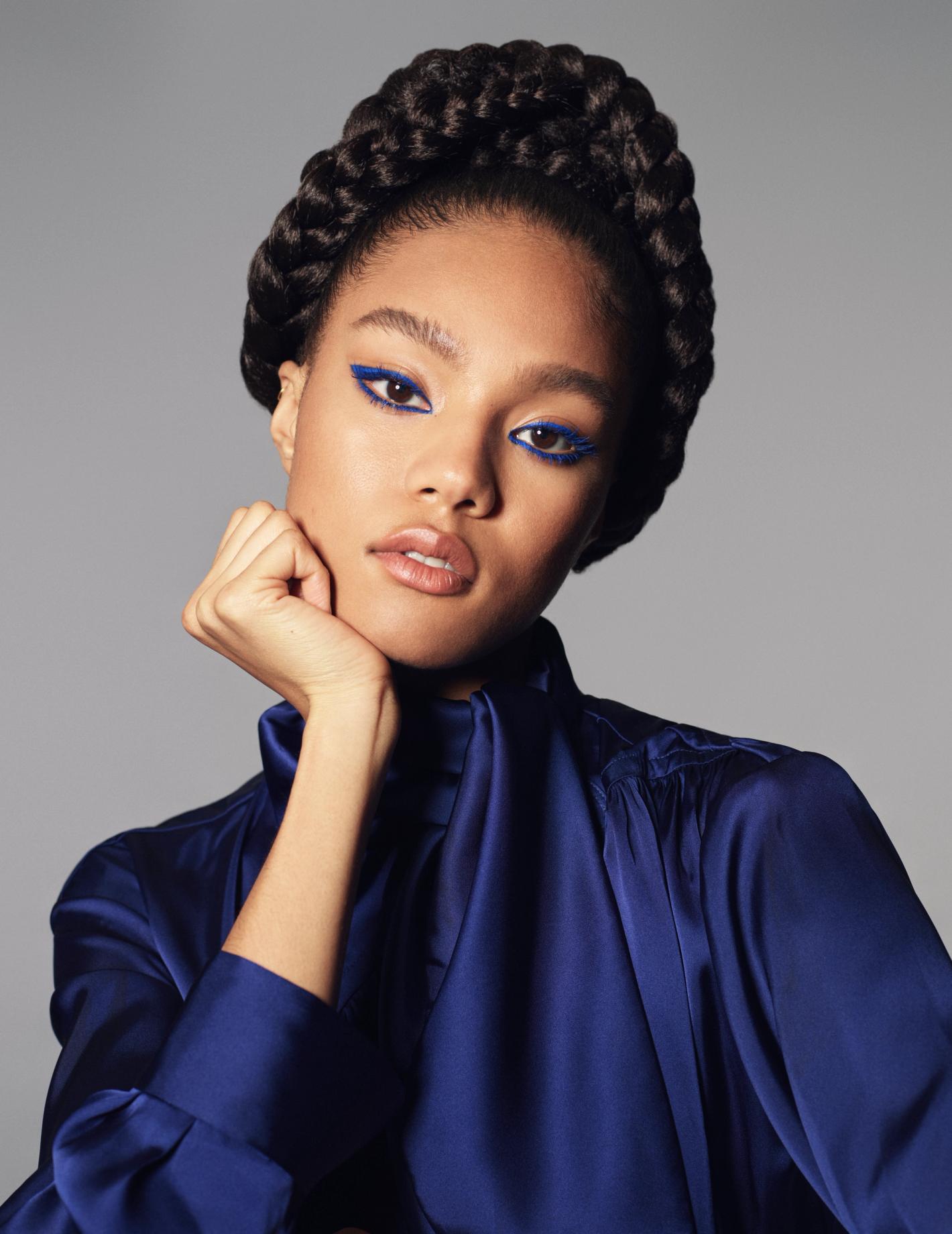 Close-up of a young woman with braided hair and bright blue eyeliner posing in a photo studio wearing a blue silk blouse.