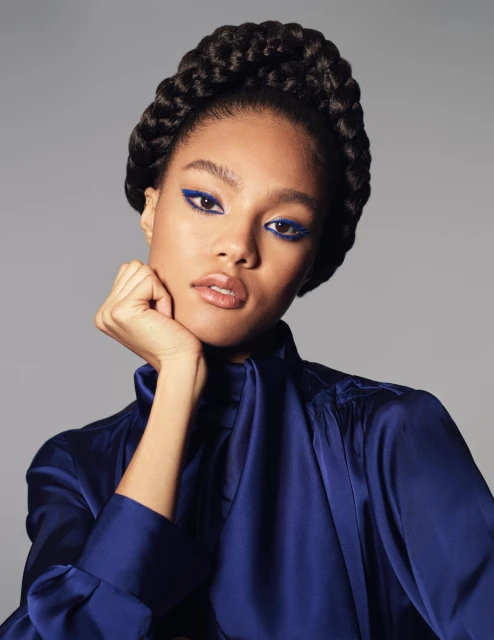 Close-up of a young woman with braided hair and bright blue eyeliner posing in a photo studio wearing a blue silk blouse.