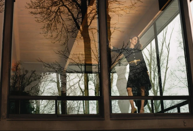 Woman in dark dress and metallic belt posed behind large glass windows reflecting trees and architecture
