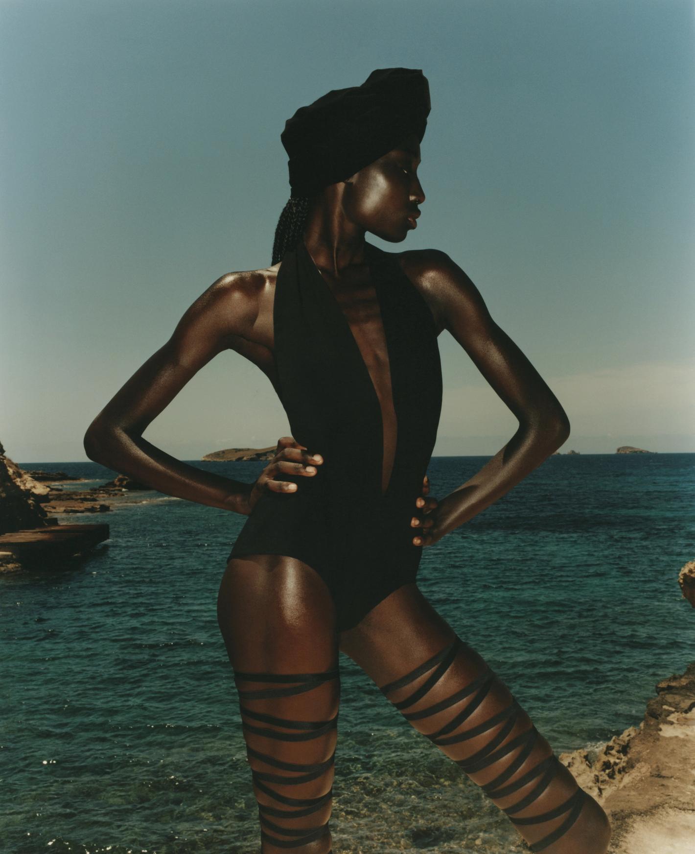 Woman in black swimsuit and headwrap stands on rock formation near ocean with island in background during a daytime outdoor shoot.