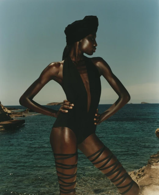 Woman in black swimsuit and headwrap stands on rock formation near ocean with island in background during a daytime outdoor shoot.